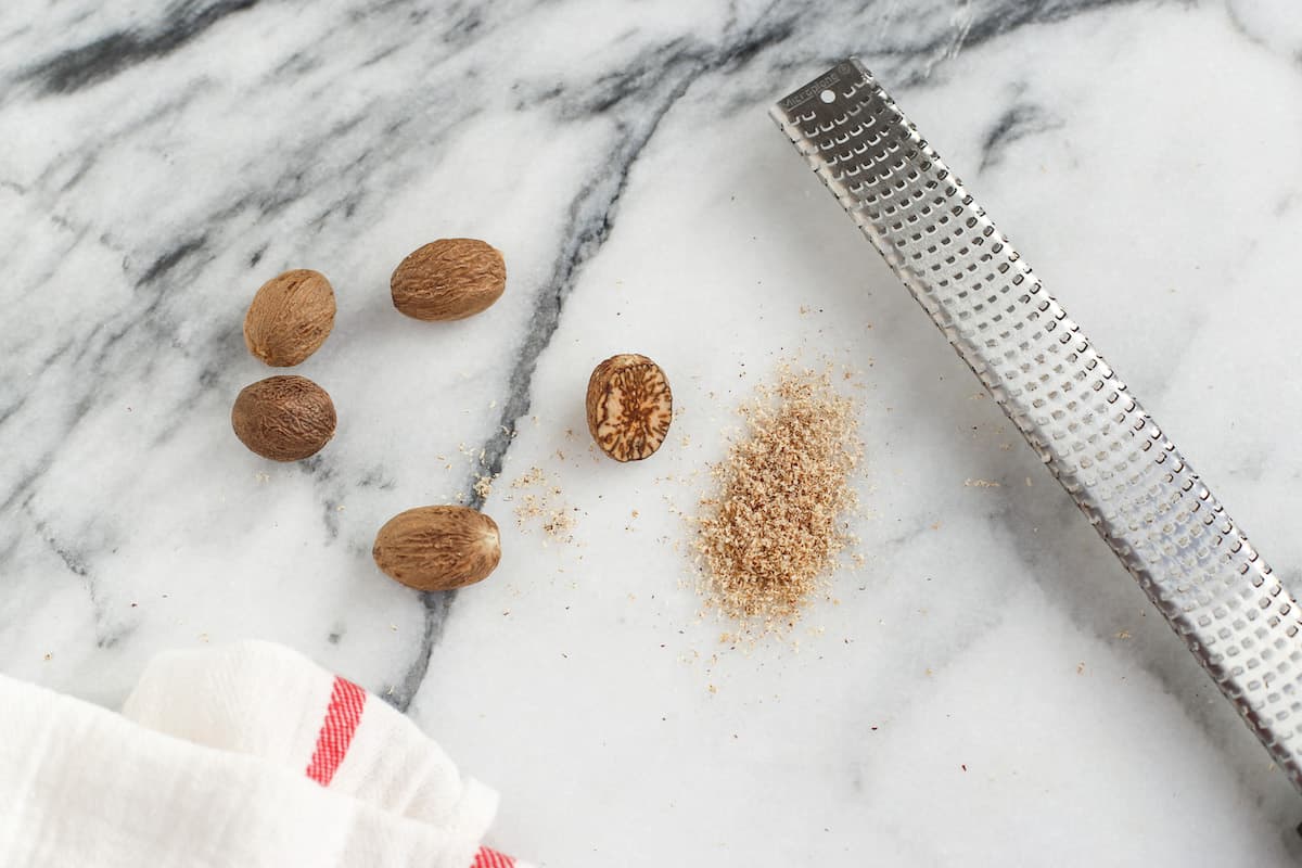 Whole nutmeg next to freshly grated nutmeg and a metal grater on a marble countertop.