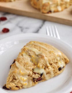 A cranberry orange scone drizzled with glaze on a white plate, with more scones on a wooden cutting board in the background.