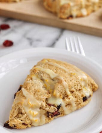A cranberry orange scone drizzled with glaze on a white plate, with more scones on a wooden cutting board in the background.