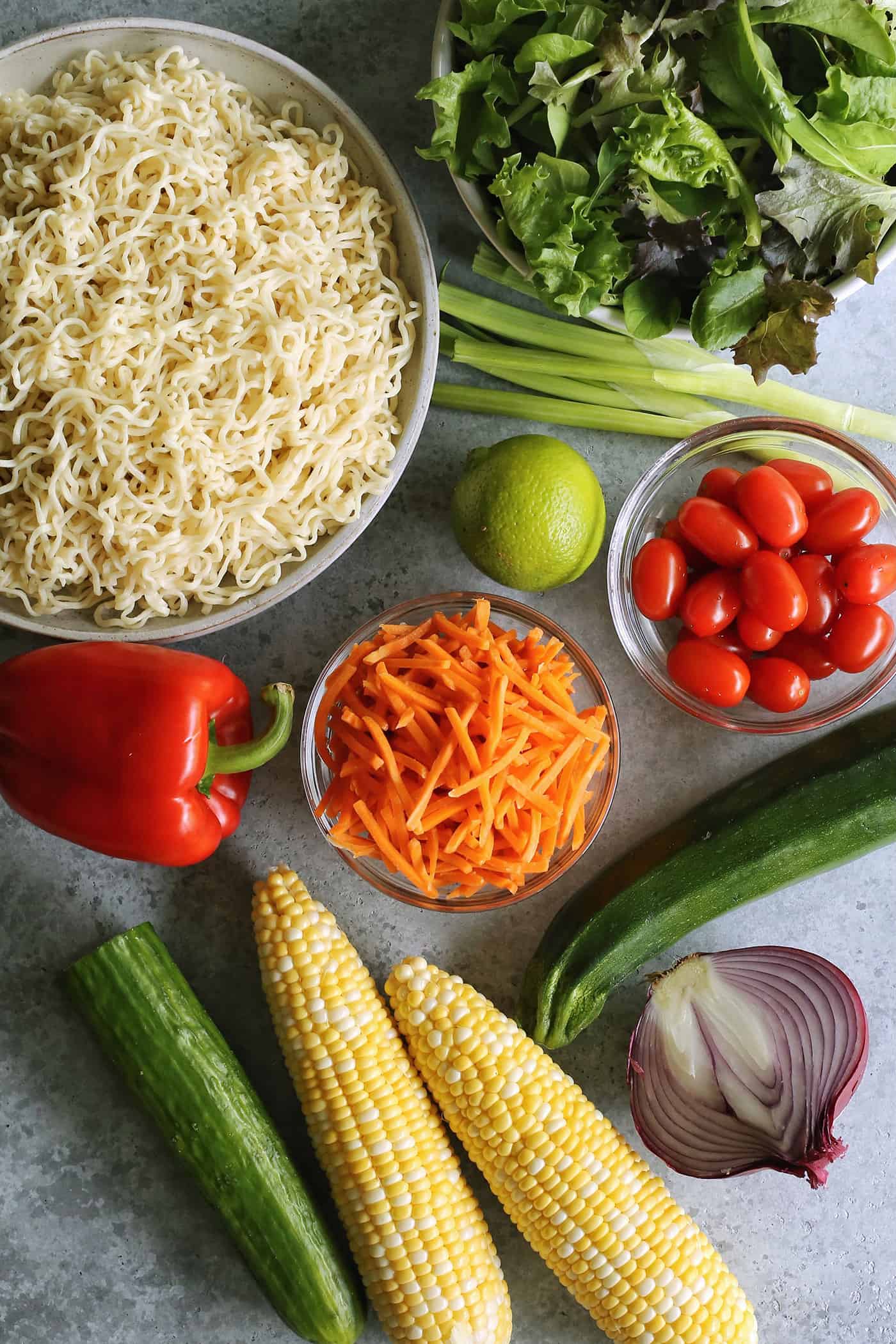 a bowl of cooked ramen noodles, plus fresh lettuce, green onion, red onion, tomatoes, lime, carrots, zucchini, sweetcorn, cucumber, and red pepper