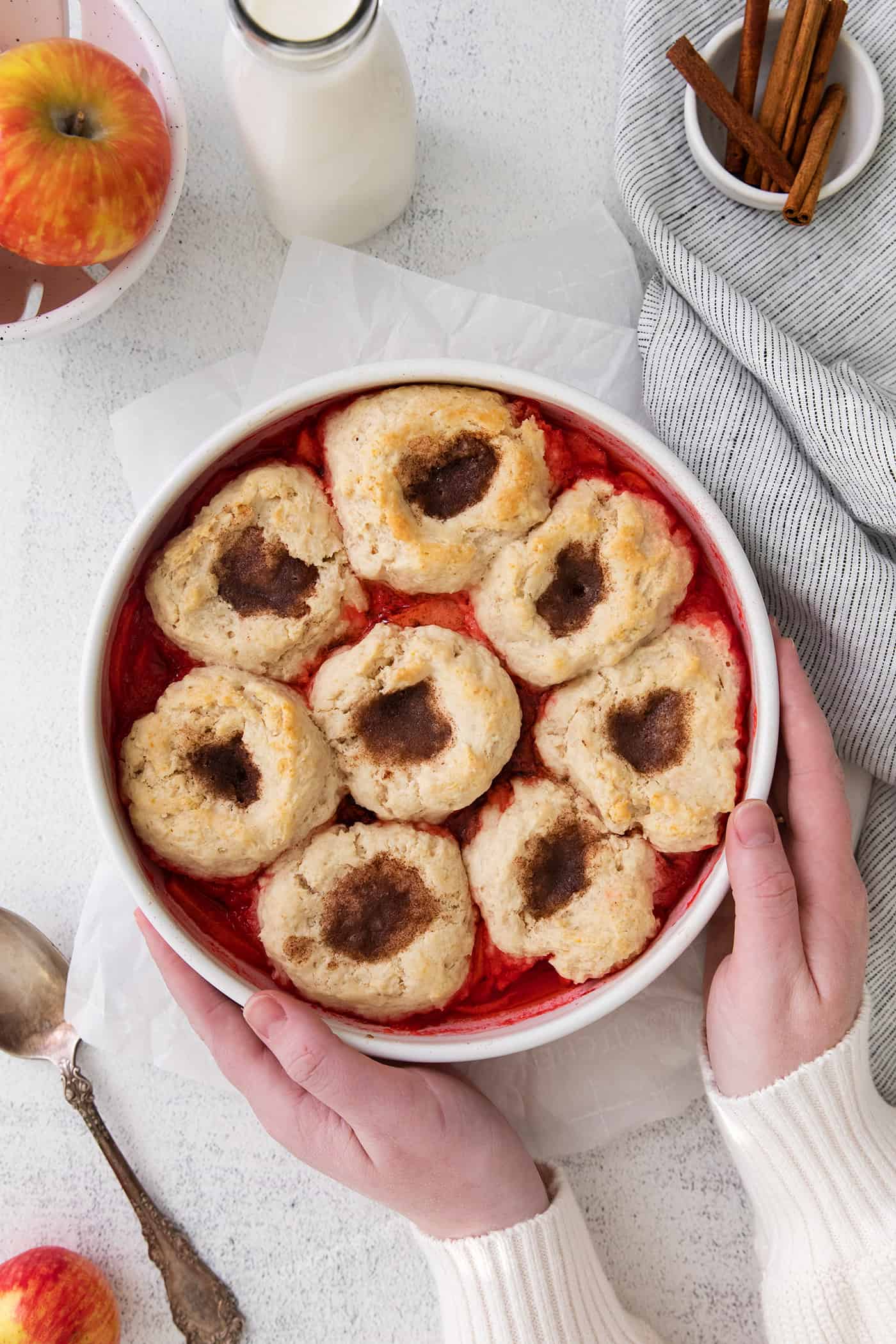 two hands holding a round white baking dish of baked apple dessert