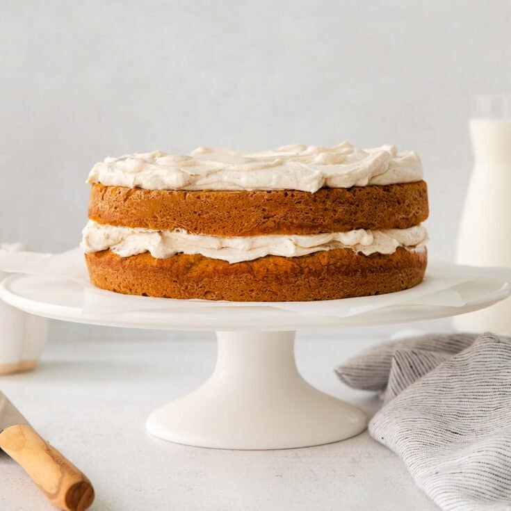 a pumpkin flavored double layer cake with creamy cinnamon whipped topping on a white cake stand