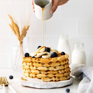 SYrup being poured over a stack of classic waffles topped with blueberries and bananas
