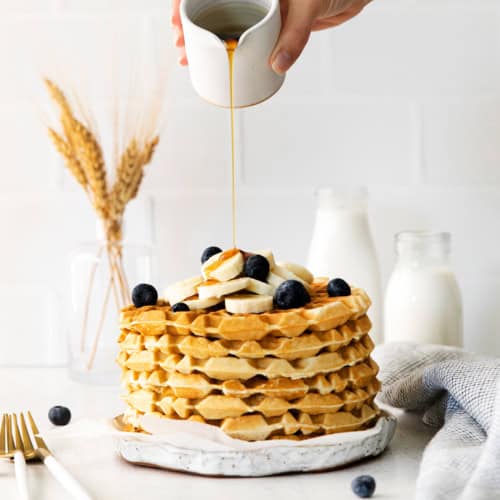 SYrup being poured over a stack of classic waffles topped with blueberries and bananas