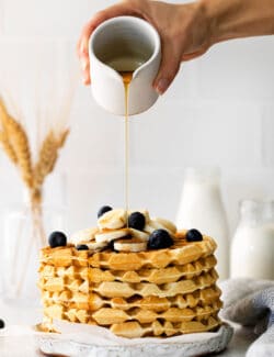 A hand drizzling maple syrup over a stack of waffles