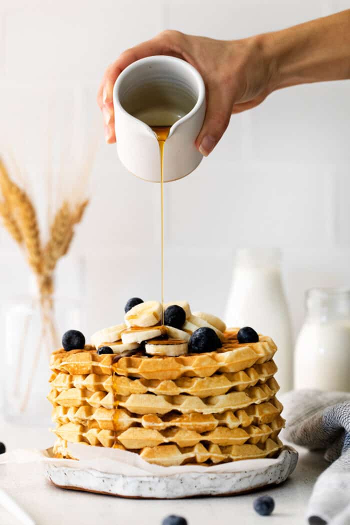 A hand drizzling maple syrup over a stack of waffles