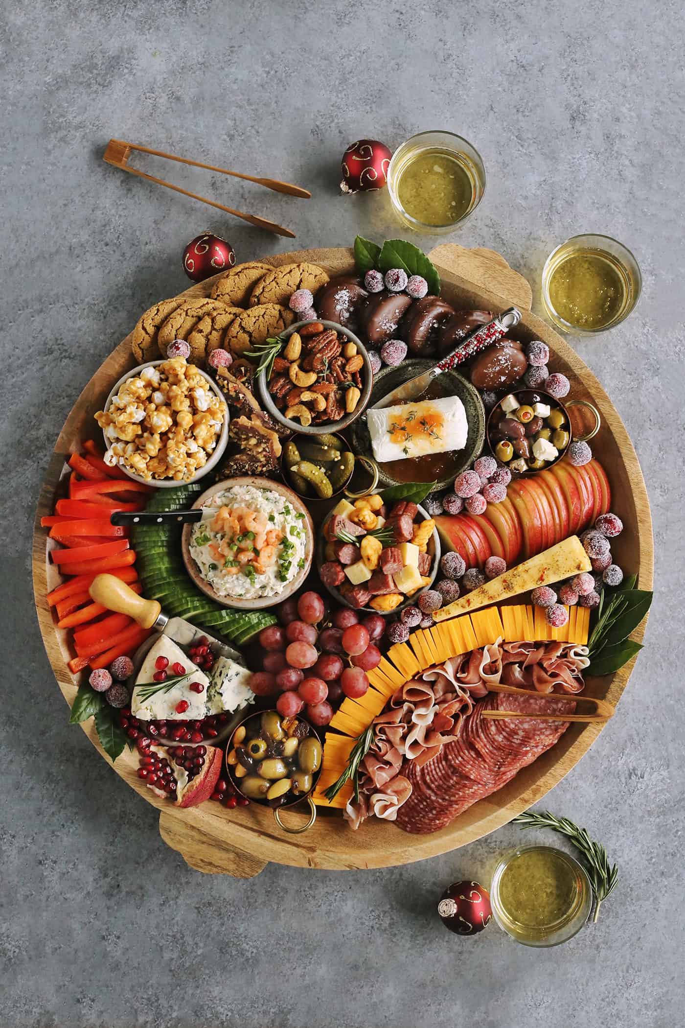 overhead photo of large round wood serving board full of charcuterie board nibbles, with glasses of white wine