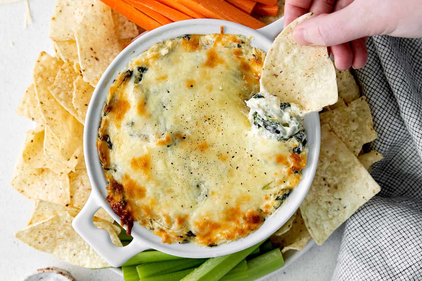 A hand dipping a tortilla chip into a dish of spinach artichoke dip