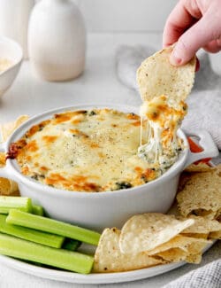 A hand dipping a chip into a dish of spinach artichoke dip
