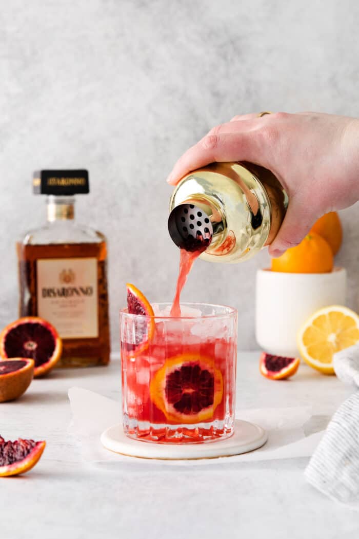 pouring the finished drink from a cocktail shaker into a cocktail glass with ice and slices of blood orange