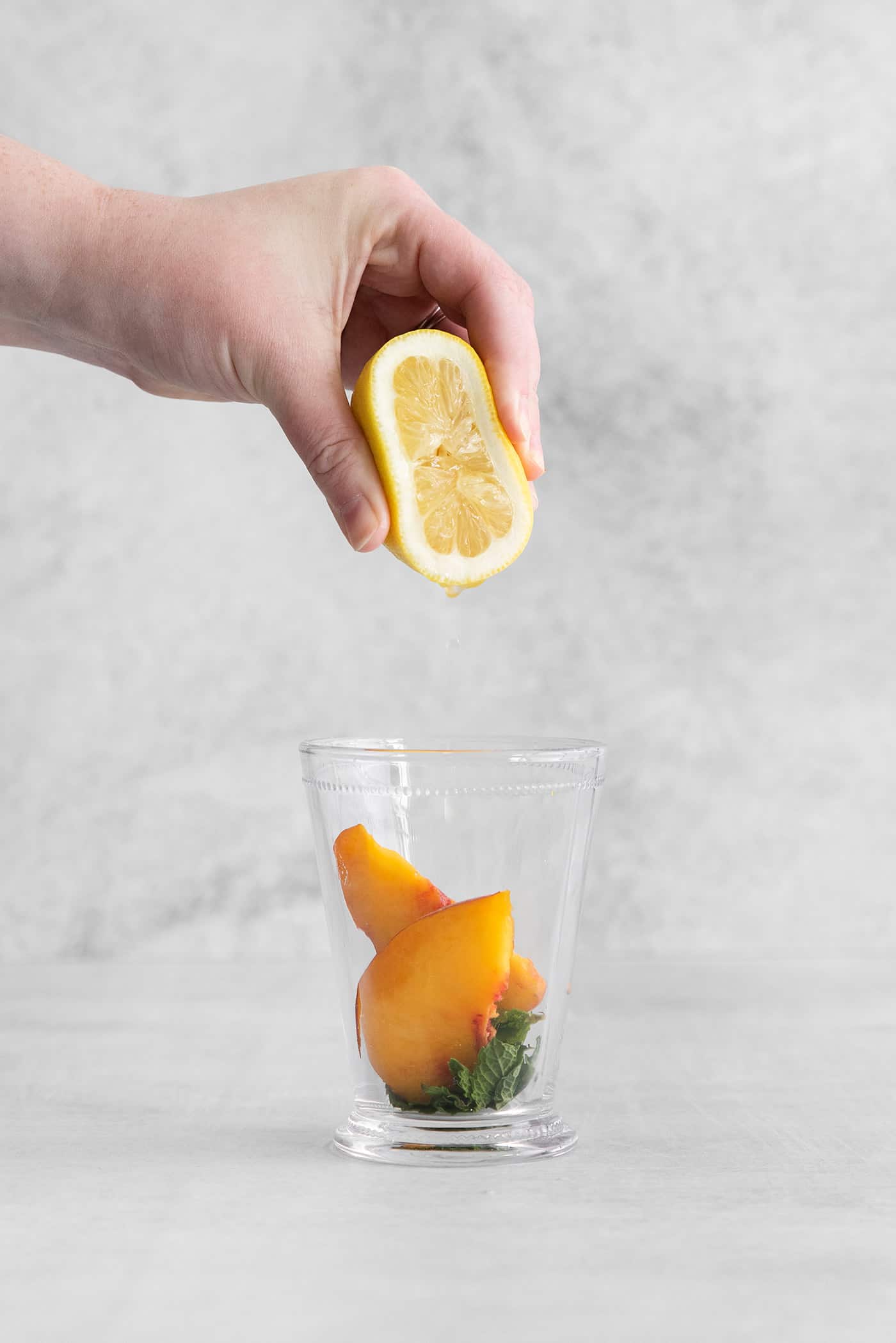 Lemon juice being squeezed into a cocktail glass with peach and mint