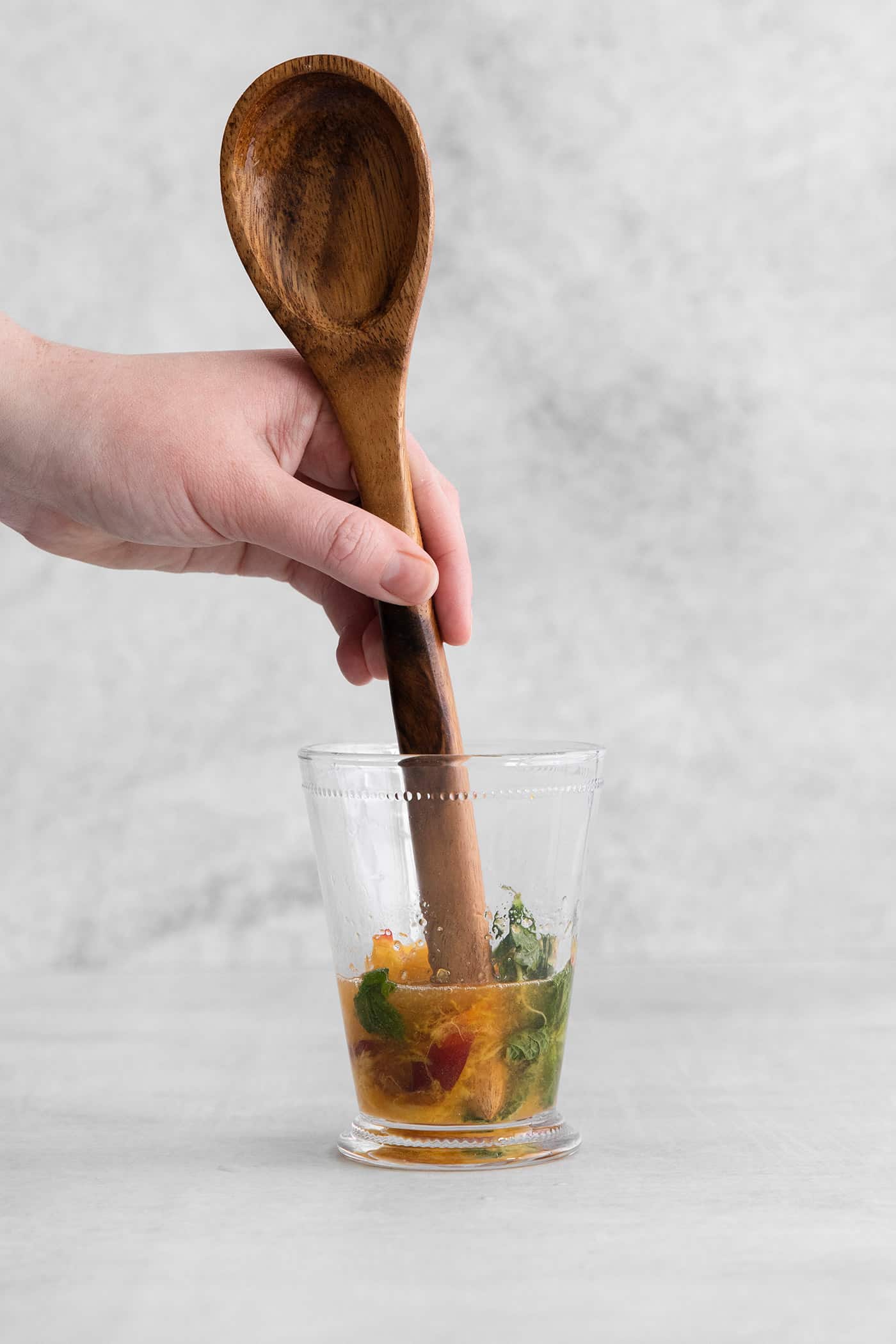 The end of a wooden spoon muddling peaches and mint in a cocktail glass