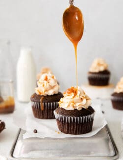 a spoon drizzling caramel sauce over chocolate cupcakes with frosting