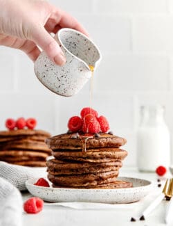 A hand pouring syrup over a stack of chocolate pancakes