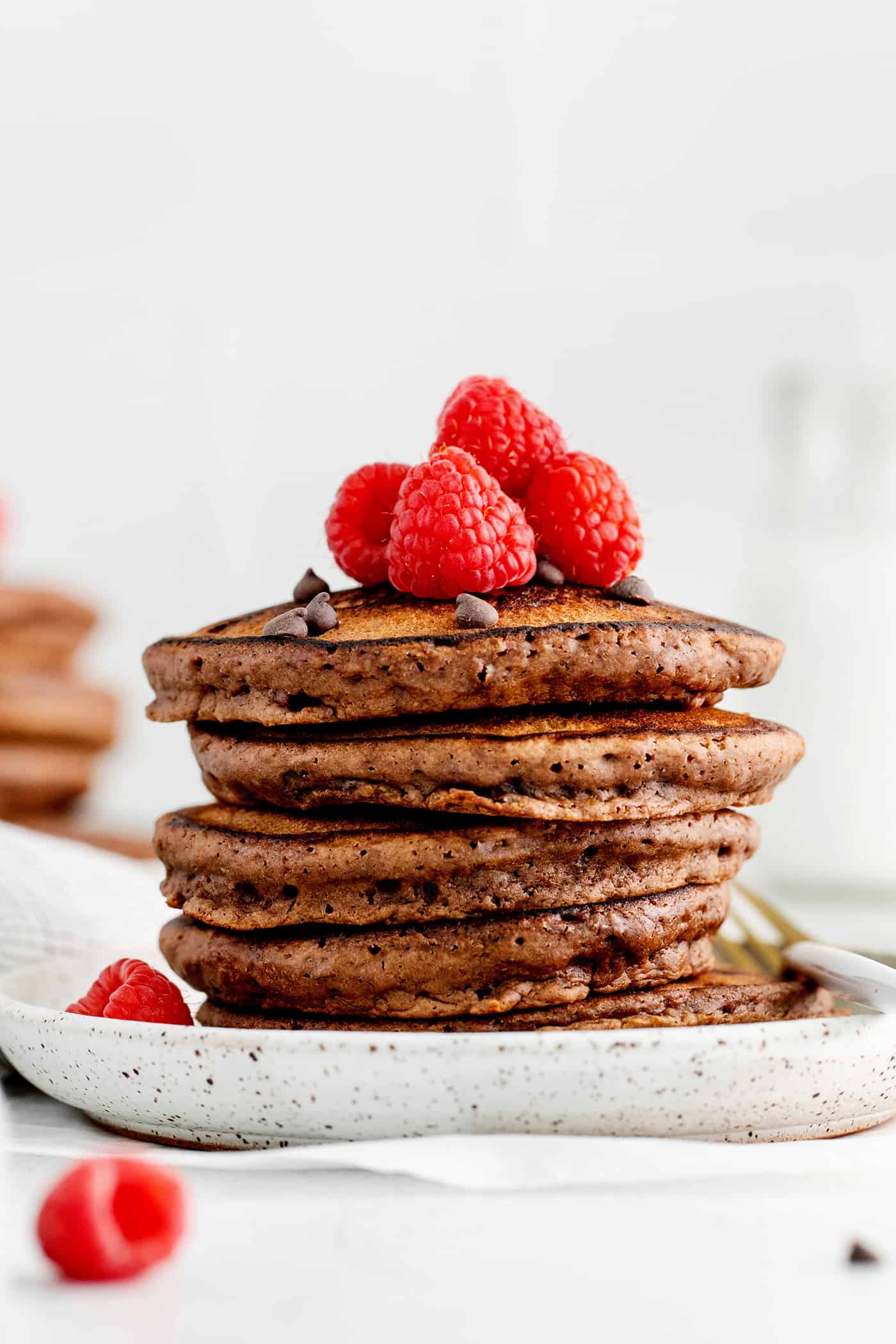 A stack of chocolate pancakes topped with raspberries on a white plate