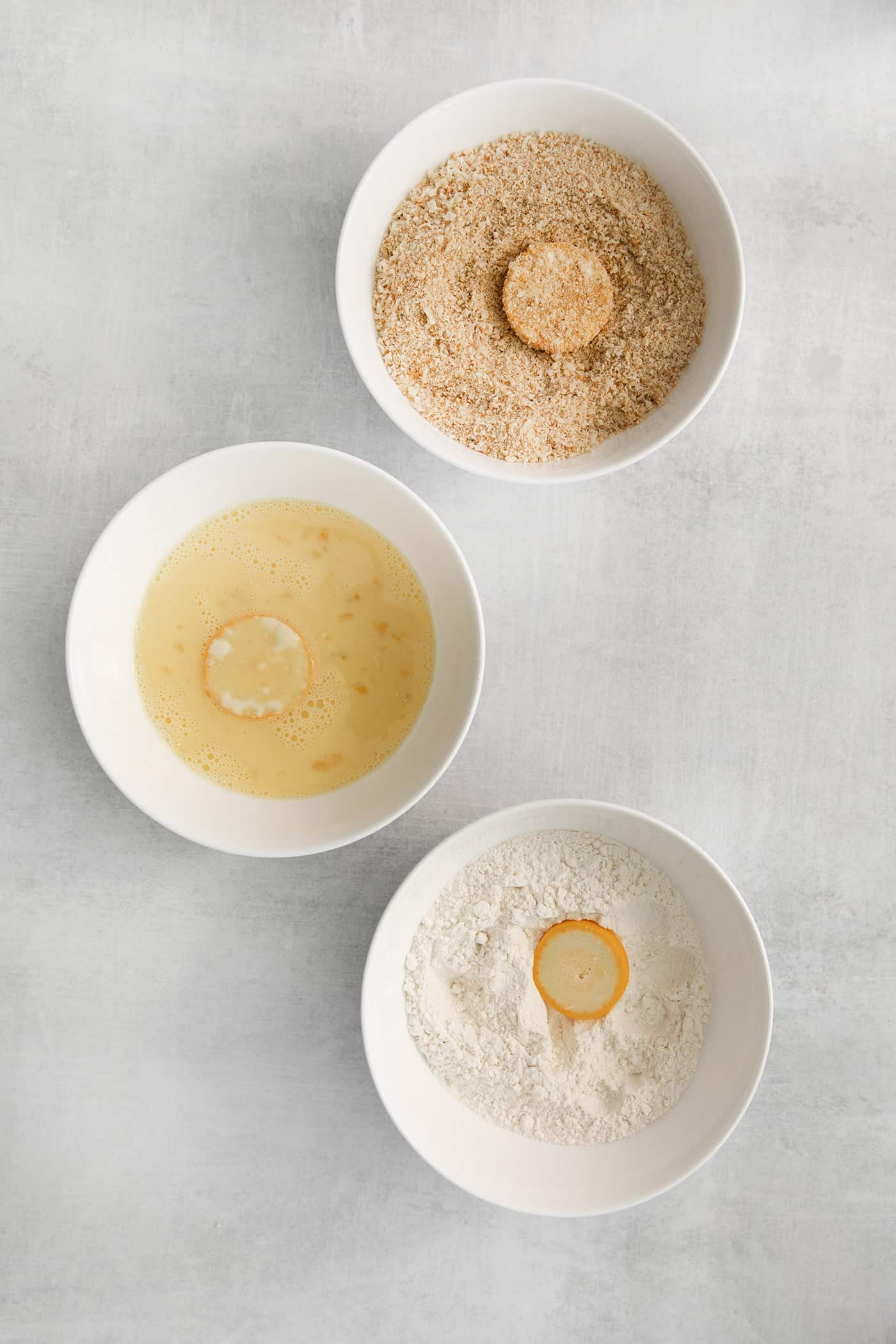 A bowl of flour, one with egg wash, and a third with breadcrumbs