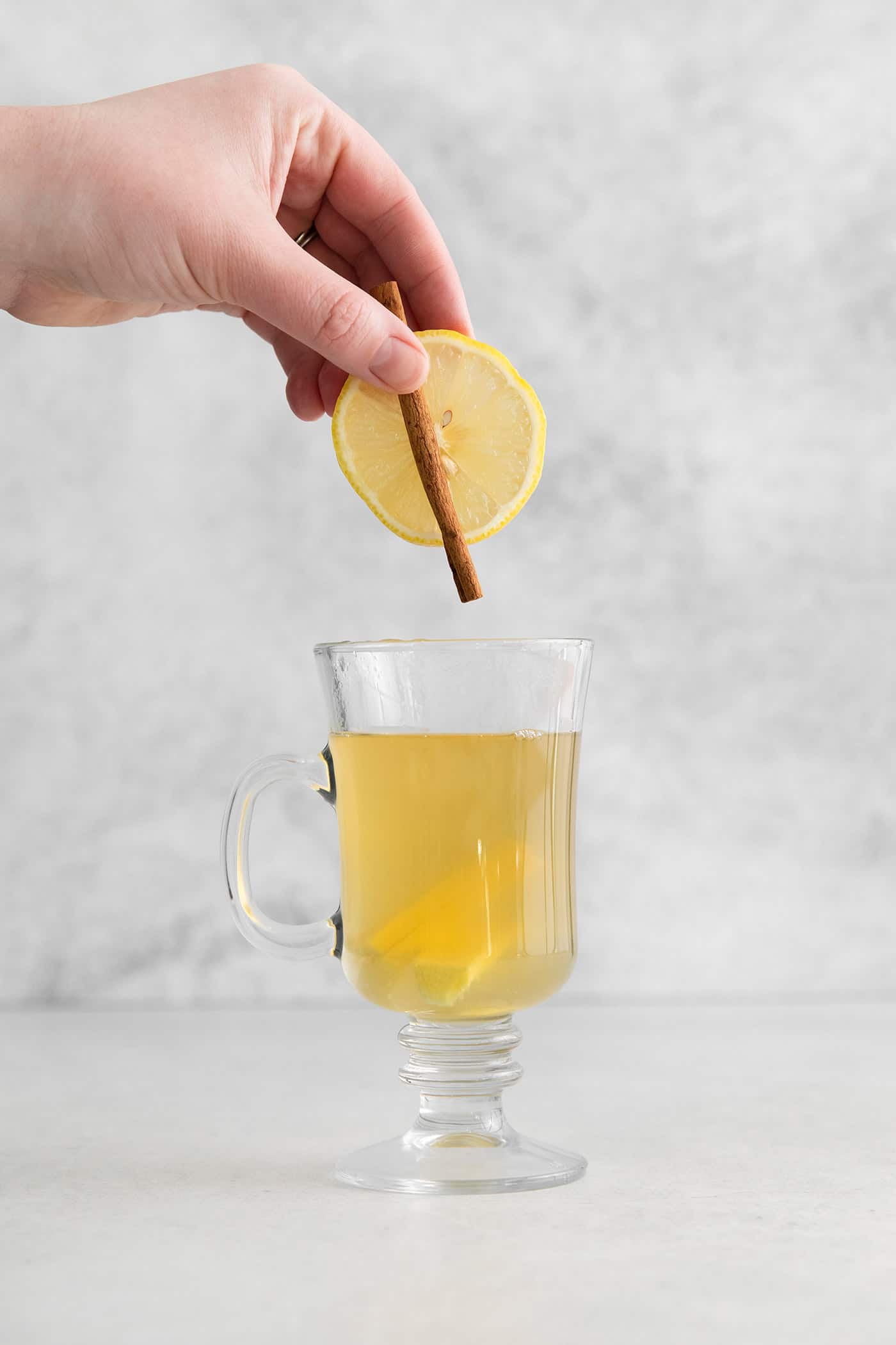 adding lemon slice and cinnamon stick to a glass of hot water with ginger