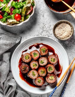 overhead photo of beef negimaki on a white plate, plus a bowl of salad