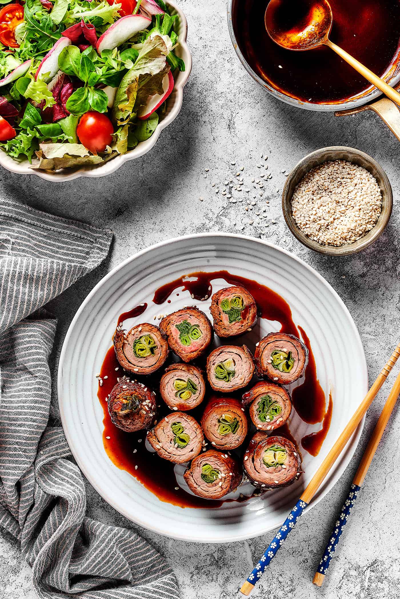 overhead photo of beef negimaki on a white plate, plus a bowl of salad