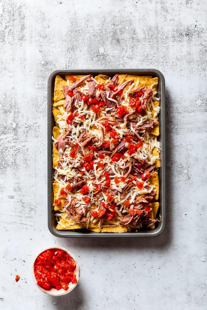 A baking sheet of tortilla chips topped with pulled pork, shredded cheese, and diced tomatoes