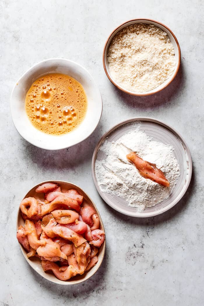 A plate of seasoned chicken strips, a bowl of flour, a bowl of breadcrumbs, and egg wash