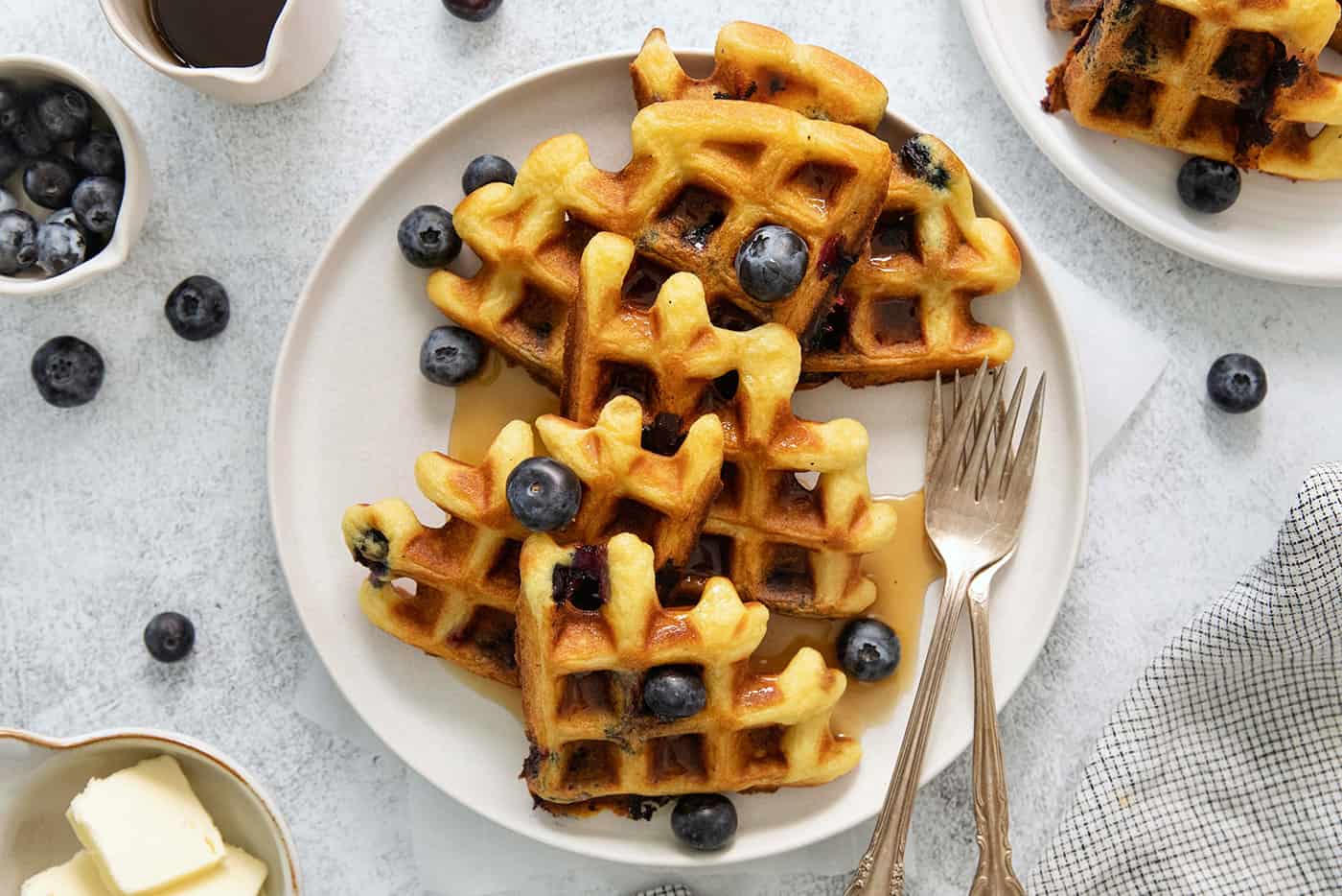 Blueberry waffles topped with blueberries are shown on a white plate with forks.