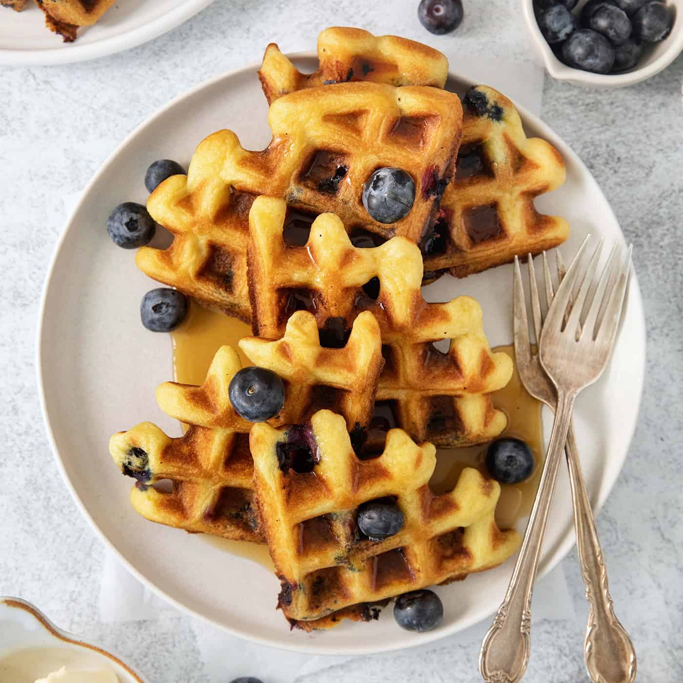 Blueberry waffles topped with blueberries are shown on a white plate with forks.
