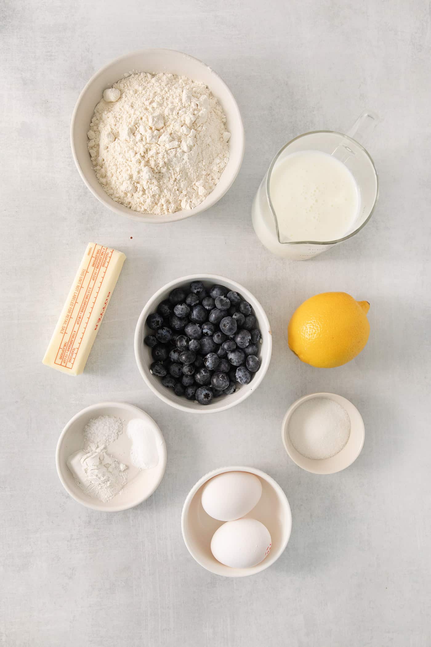 An overview of ingredients needed to make blueberry waffles including butter, flour, sugar, lemon, milk, and blueberries.
