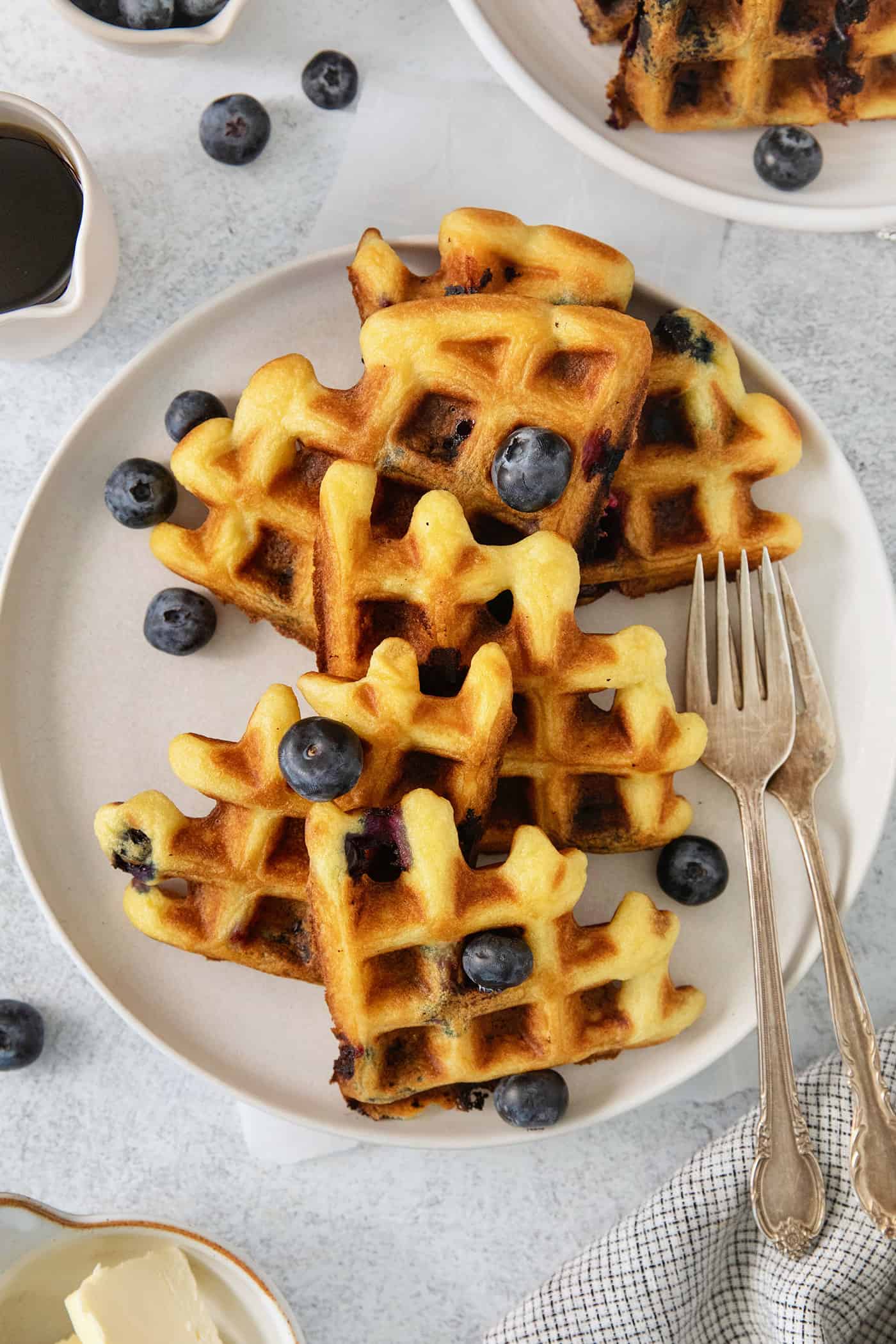Blueberry waffles topped with blueberries are shown on a white plate with forks.
