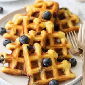 Blueberry waffles topped with blueberries are shown on a white plate with forks.