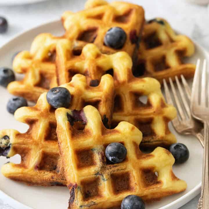 Blueberry waffles topped with blueberries are shown on a white plate with forks.