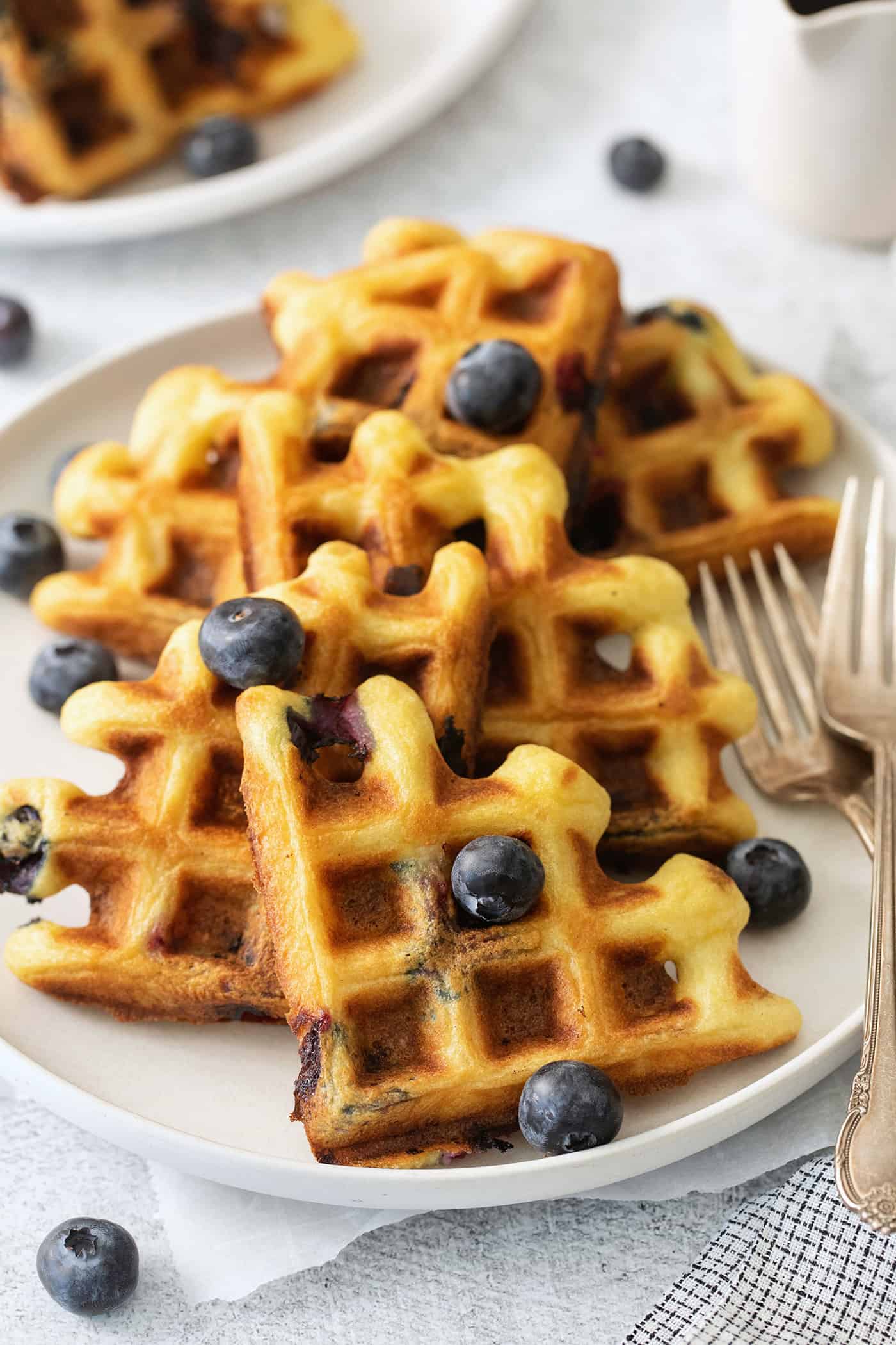 Blueberry waffles topped with blueberries are shown on a white plate with forks.