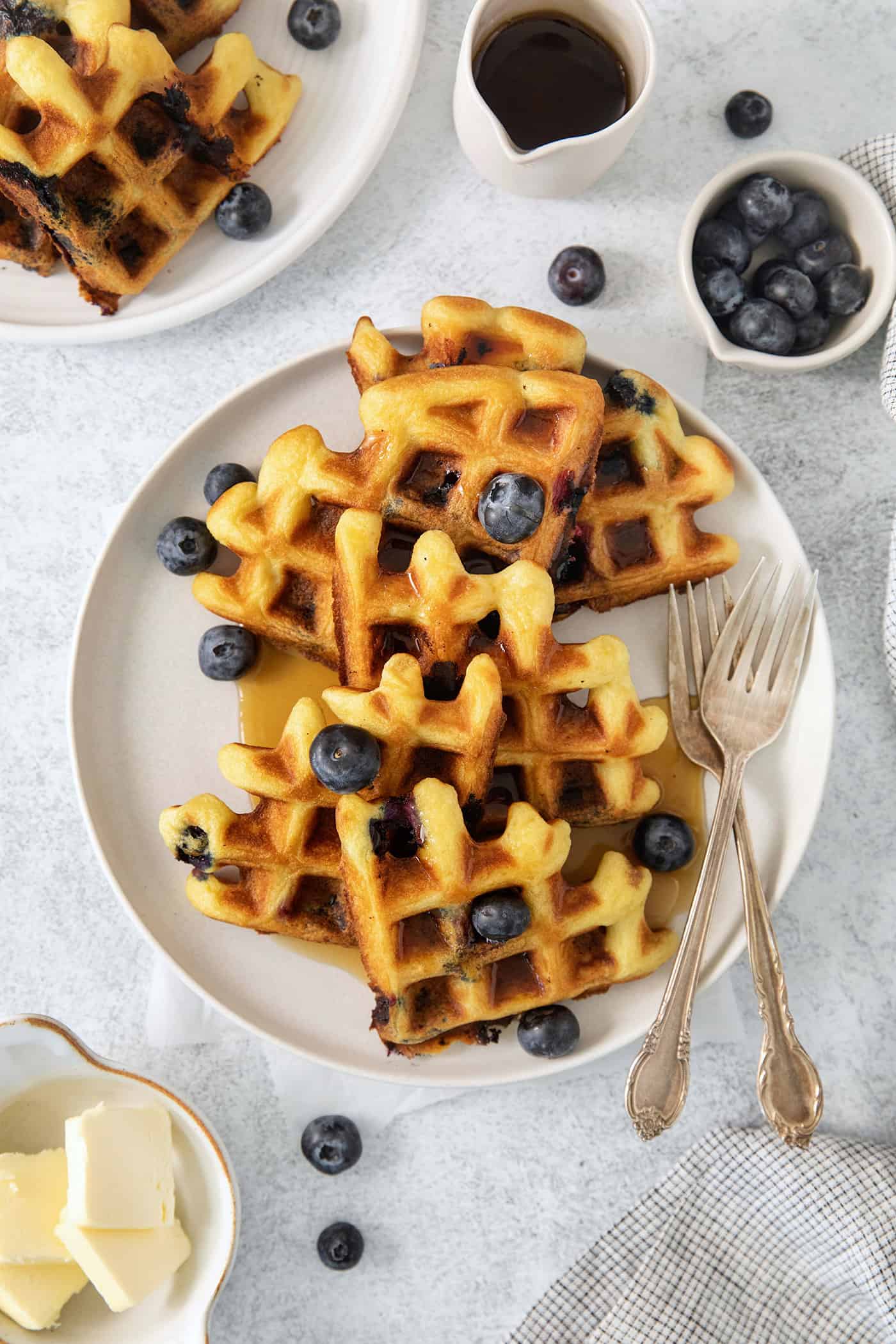 Blueberry waffles topped with blueberries are shown on a white plate with forks.