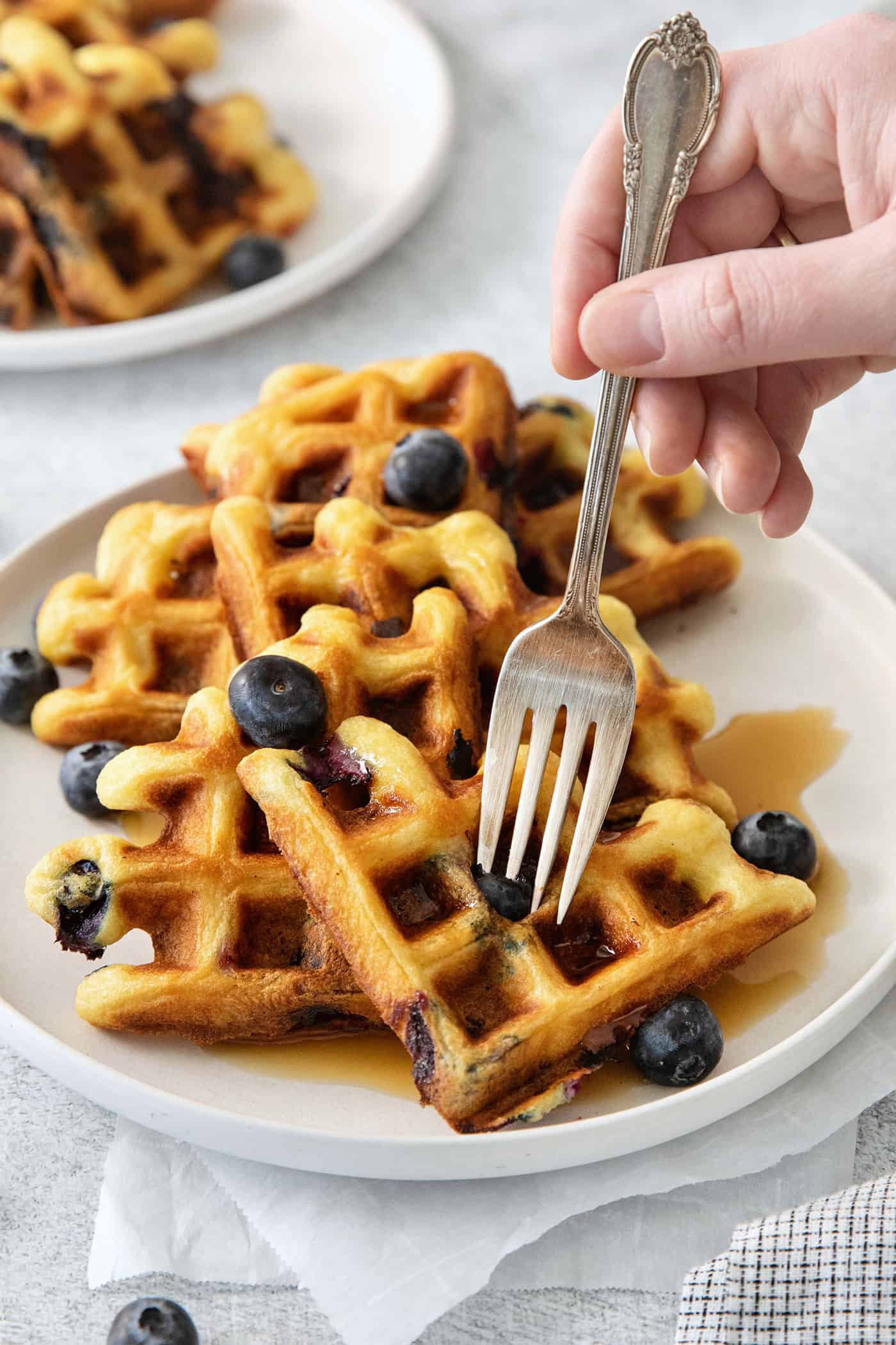 A hand holds a fork to cup into a plate of blueberry waffles.