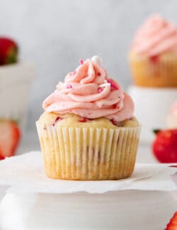 A frosted strawberry cupcake is show on a cupcake stand with cupcakes and strawberries in the background.