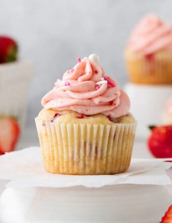 A frosted strawberry cupcake is show on a cupcake stand with cupcakes and strawberries in the background.