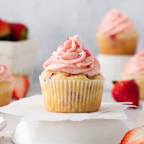 A frosted strawberry cupcake is show on a cupcake stand with cupcakes and strawberries in the background.