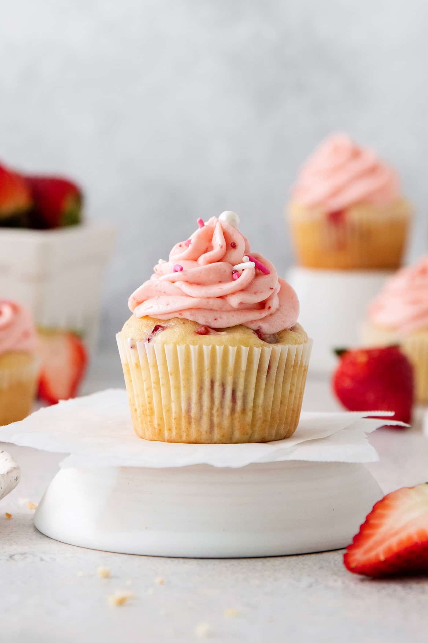 A frosted strawberry cupcake is show on a cupcake stand with cupcakes and strawberries in the background.