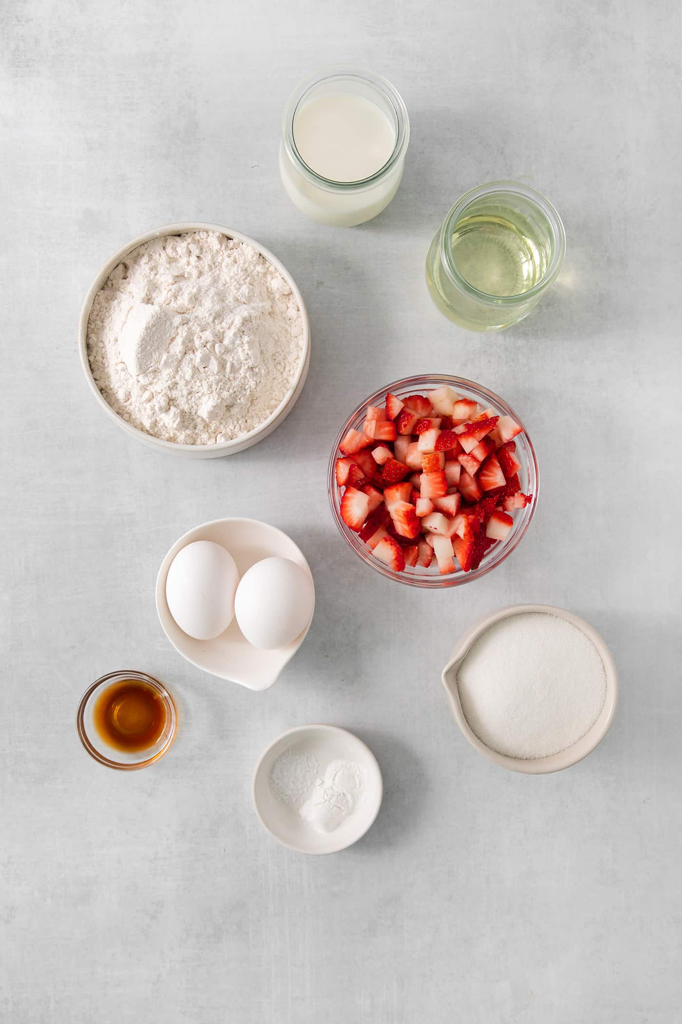 Ingredients needed to make strawberry cupcakes are shown, including fresh strawberries, vanilla extract, eggs, flour, sugar, and oil.