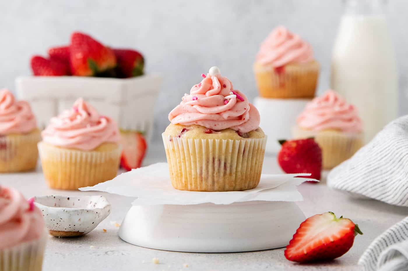 A focused shot of a strawberry cupcake with pink frosting with cupcakes and strawberries in the background.