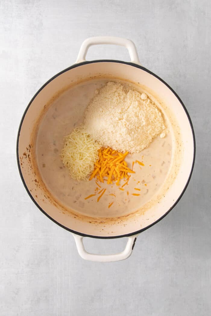 Mozzarella, cheddar, and parmesan cheeses are added to cream to make a sauce in a pot.