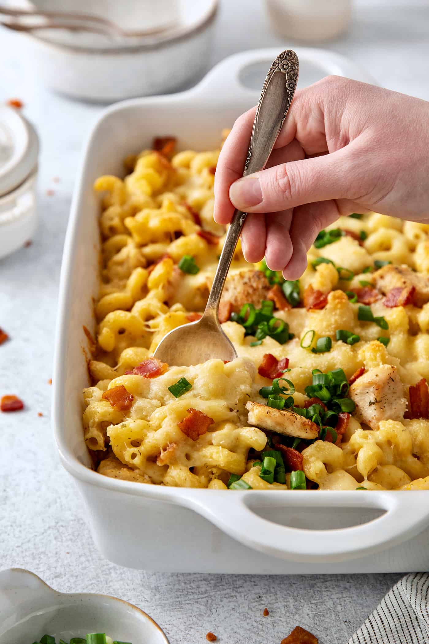 This Chicken Bacon Ranch Pasta is a perfect family-friendly meal loaded with flavor! Bacon, chicken, and pasta are baked in a creamy sauce. A hand holding a spoon digs into a casserole dish of chicken bacon ranch pasta.