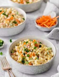 Bowls of hawaiian macaroni salad are seen on a table with shredded carrots in the background.