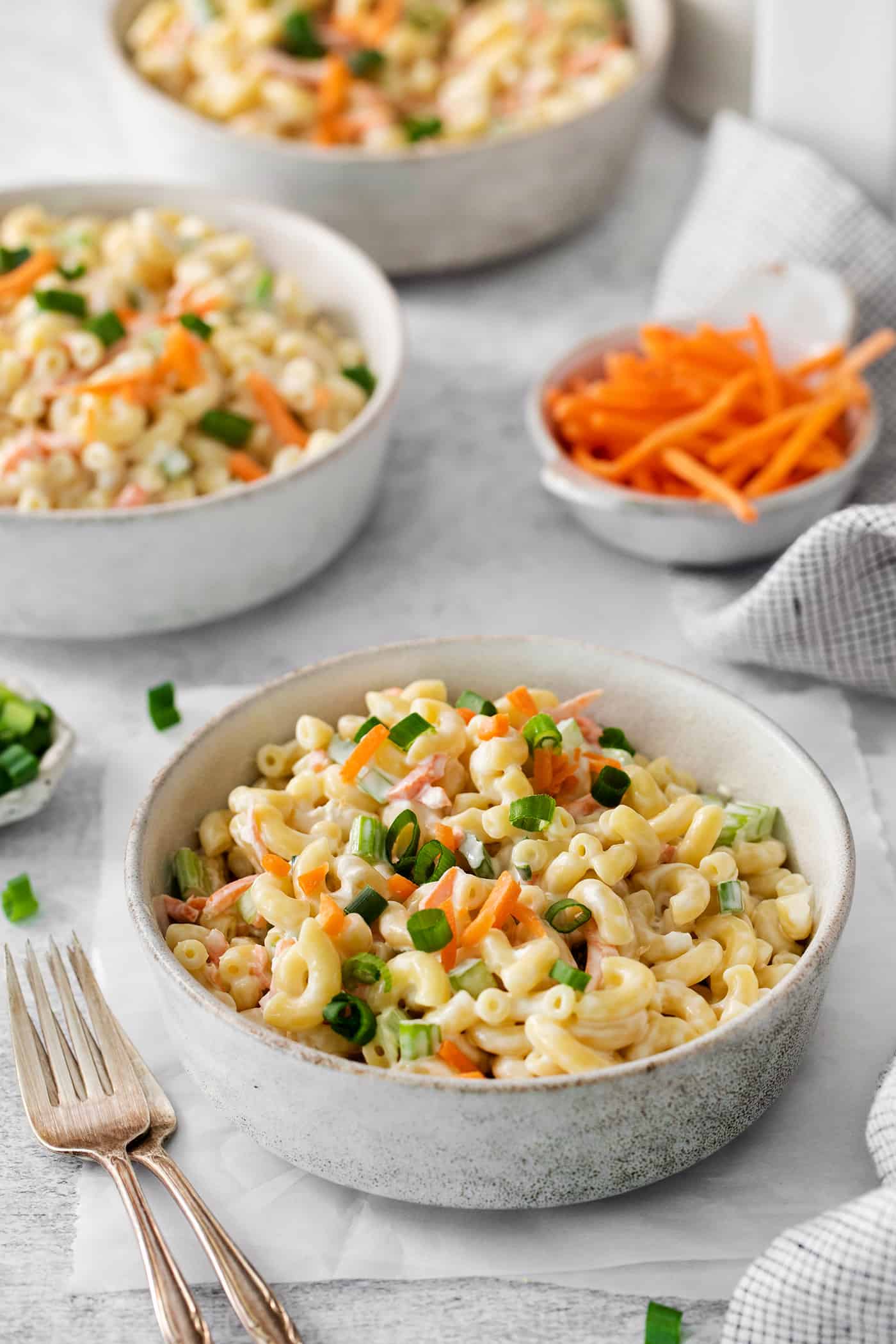 Bowls of hawaiian macaroni salad are seen on a table with shredded carrots in the background.
