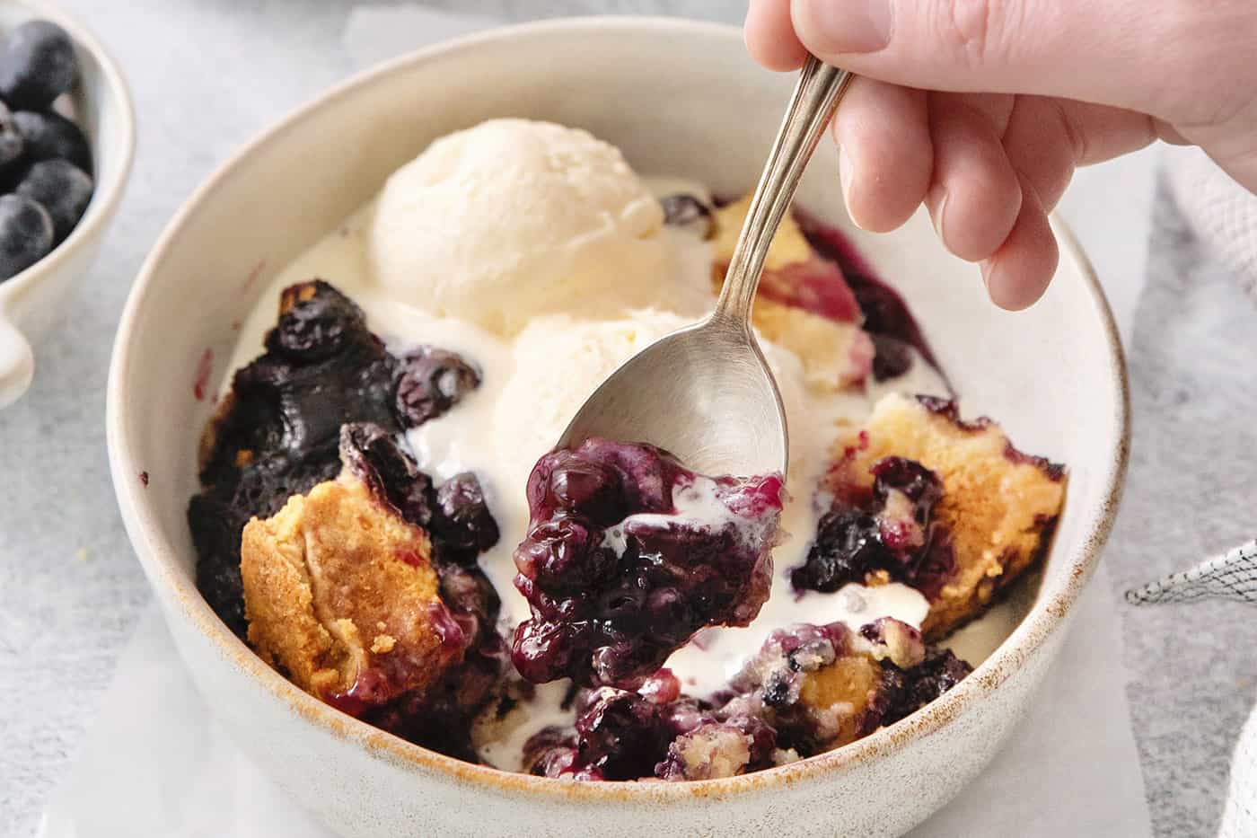 A hand holds a spoon dipping into a bowl of blueberry dump cake.