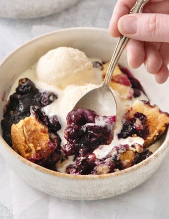 A hand holds a spoon dipping into a bowl of blueberry dump cake.