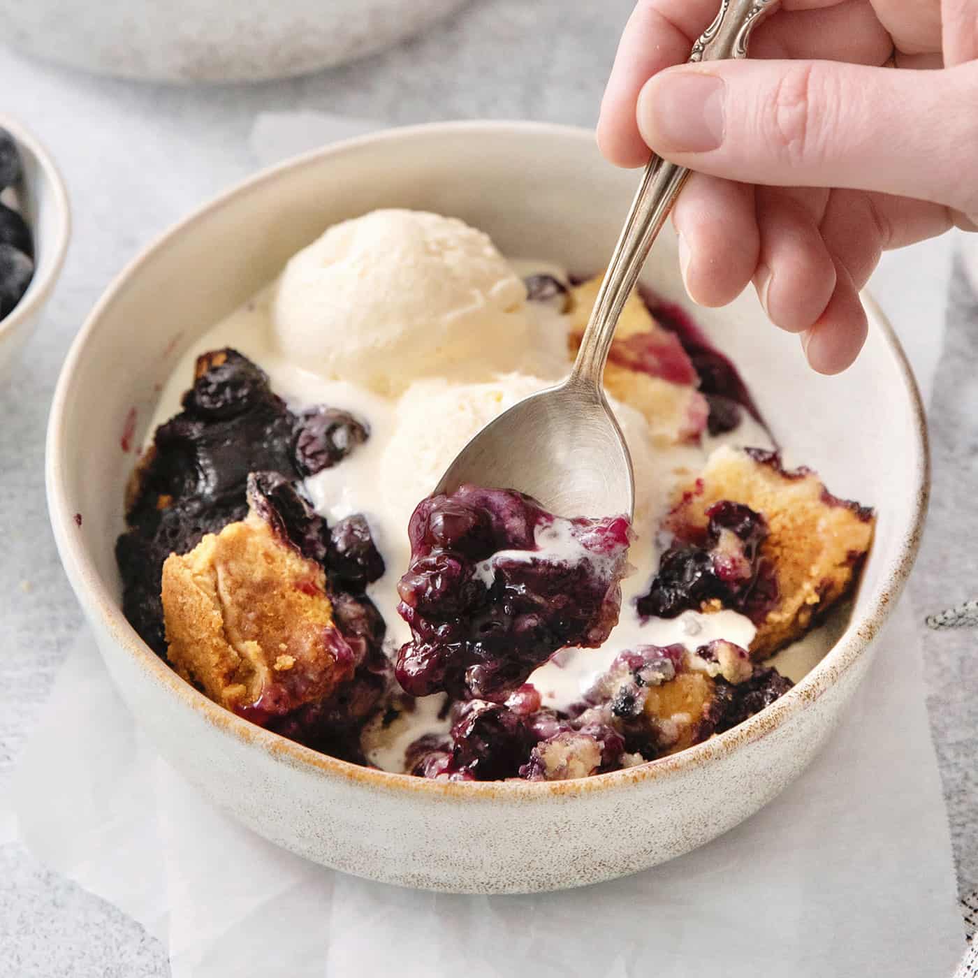 A hand holds a spoon dipping into a bowl of blueberry dump cake.