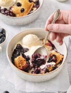 A hand holds a spoon dipping into a bowl of blueberry dump cake.