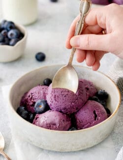 Blueberry ice cream in a bowl with a hand and a spoon.