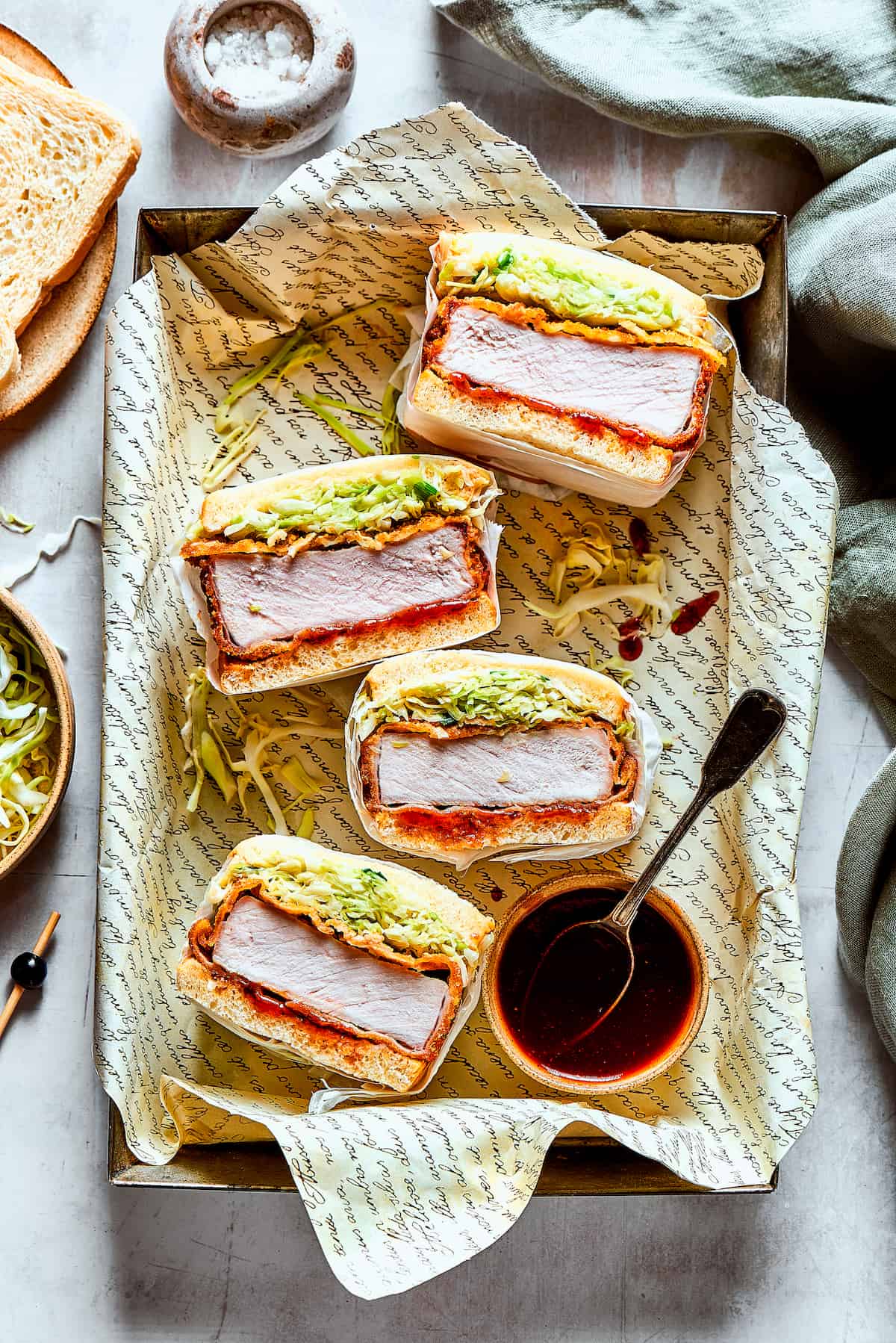Katsu Sando is a Japanese classic, with a crispy pork cutlet, fresh, shredded cabbage, and a drizzle of homemade sauce on white bread. A stack of katsu sandos and tonkatsu sauce.